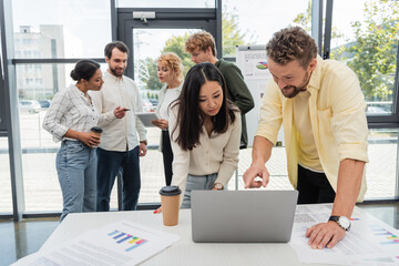 positive businessman pointing at laptop near asian colleague while interracial team talking on background