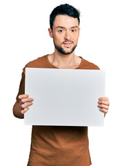 Hispanic man with beard holding blank empty banner relaxed with serious expression on face. simple and natural looking at the camera.