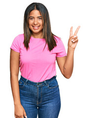 Young latin girl wearing casual clothes smiling with happy face winking at the camera doing victory sign. number two.