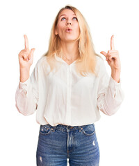 Young beautiful blonde woman wearing casual shirt amazed and surprised looking up and pointing with fingers and raised arms.