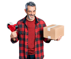 Middle age hispanic with grey hair holding packing tape and cardboard box winking looking at the...