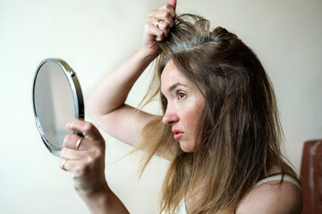 Young woman looks in the mirror at her gray hair roots. Problem of gray hair and loss hair