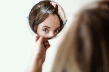 Close-up of a mirror with a reflection of a woman's face. Young woman is examining her gray hair...