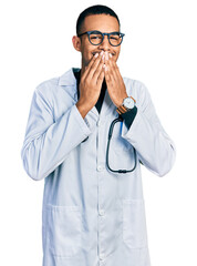 Young african american man wearing doctor uniform and stethoscope laughing and embarrassed giggle covering mouth with hands, gossip and scandal concept