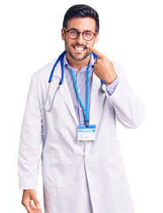 Young hispanic man wearing doctor uniform and stethoscope pointing with hand finger to face and nose, smiling cheerful. beauty concept