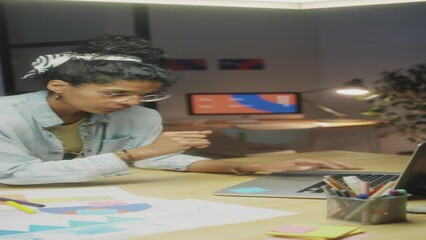 Vertical shot of Latin businesswoman in casual outfit leaning on desk, using laptop and writing down on sticky notes while working in office during night - Powered by Adobe