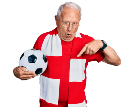 Senior man with grey hair football hooligan holding ball pointing down with fingers showing advertisement, surprised face and open mouth