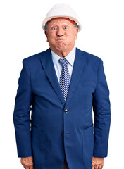 Senior handsome grey-haired man wearing suit and architect hardhat puffing cheeks with funny face. mouth inflated with air, crazy expression.