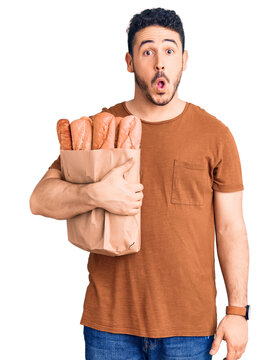 Young hispanic man holding paper bag with bread scared and amazed with open mouth for surprise, disbelief face