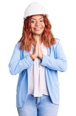 Young latin woman wearing architect hardhat praying with hands together asking for forgiveness smiling confident.