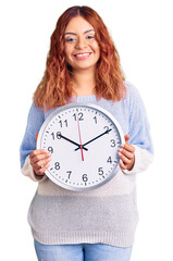 Young latin woman holding big clock looking positive and happy standing and smiling with a confident smile showing teeth