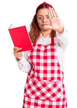 Young latin woman wearing apron holding recipe book with open hand doing stop sign with serious and confident expression, defense gesture