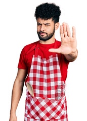 Young arab man with beard wearing cook apron doing stop sing with palm of the hand. warning expression with negative and serious gesture on the face.