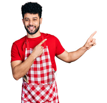 Young arab man with beard wearing cook apron smiling and looking at the camera pointing with two hands and fingers to the side.