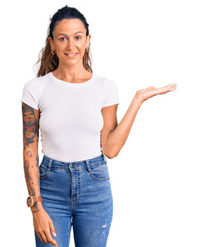 Young hispanic woman with tattoo wearing casual white tshirt smiling cheerful presenting and pointing with palm of hand looking at the camera.