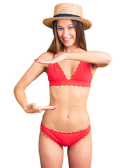 Beautiful brunette young woman wearing bikini gesturing with hands showing big and large size sign, measure symbol. smiling looking at the camera. measuring concept.