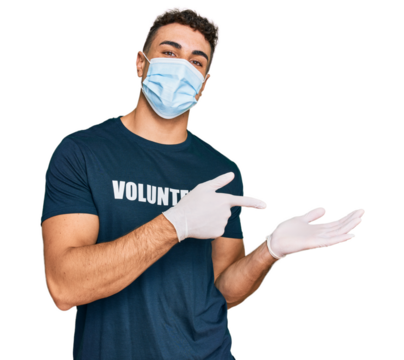 Hispanic young man wearing medical mask and volunteer t shirt amazed and smiling to the camera while presenting with hand and pointing with finger.