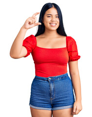 Young beautiful asian girl wearing casual clothes smiling and confident gesturing with hand doing small size sign with fingers looking and the camera. measure concept.