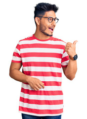 Handsome latin american young man wearing casual clothes and glasses smiling with happy face looking and pointing to the side with thumb up.