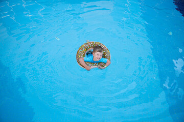A little happy boy bathes in a doughnut lifebuoy in clear water in swimming pool in sunny weather