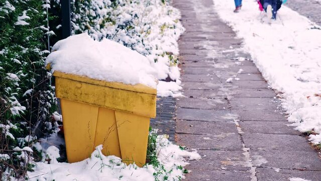 Yellow Plastic Winter Grit Sand Box Covered in Snow Standing by Icy Pavement