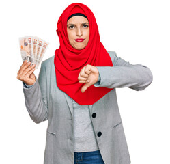 Beautiful hispanic woman wearing islamic hijab holding 10 pounds banknotes with angry face,...