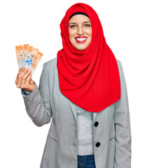 Beautiful hispanic woman wearing islamic hijab holding 10 swiss franc banknotes looking positive and happy standing and smiling with a confident smile showing teeth