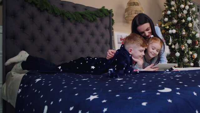 Mother and children lie on the bed and play on the tablet computer. Christmas.