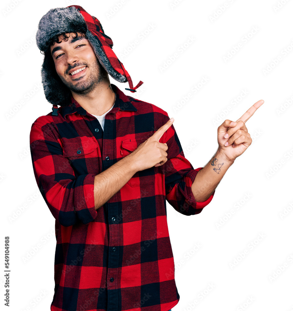 Wall mural Hispanic young man with beard wearing fluffy earmuff hat smiling and looking at the camera pointing with two hands and fingers to the side.