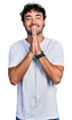 Fototapeta na wymiar Hispanic young man with beard wearing casual white t shirt praying with hands together asking for forgiveness smiling confident.