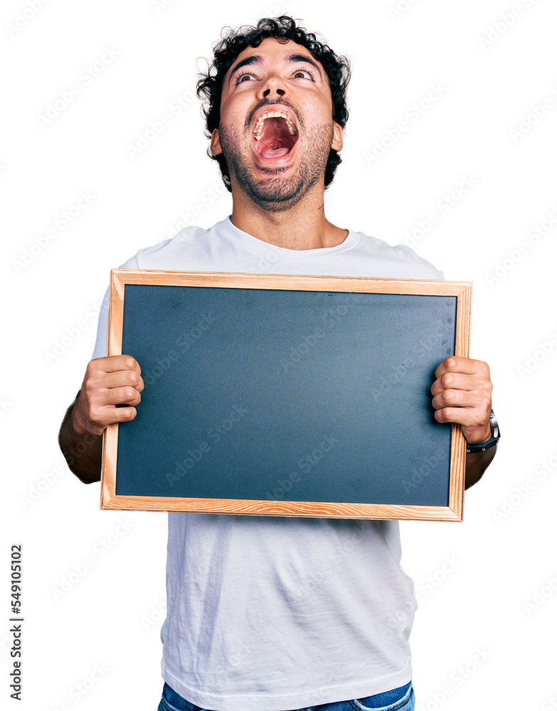 Sticker Hispanic young man with beard holding blackboard angry and mad screaming frustrated and furious, shouting with anger looking up.