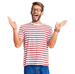 Naklejka premium Handsome blond man with beard wearing casual clothes and glasses celebrating mad and crazy for success with arms raised and closed eyes screaming excited. winner concept