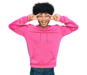 Young african american man with afro hair wearing casual pink sweatshirt doing peace symbol with fingers over face, smiling cheerful showing victory