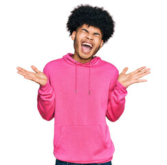 Young african american man with afro hair wearing casual pink sweatshirt celebrating mad and crazy for success with arms raised and closed eyes screaming excited. winner concept