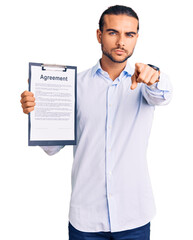 Young handsome man holding clipboard with agreement document pointing with finger to the camera and to you, confident gesture looking serious