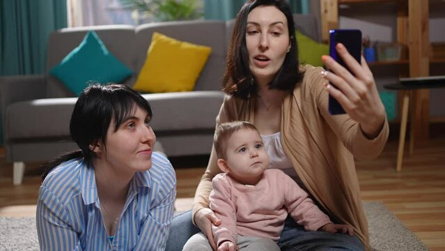 a two young mothers with a child are sitting in the living room and communicating via video communication smartphone in hand