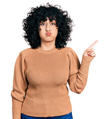 Young middle east girl pointing with finger to the side puffing cheeks with funny face. mouth inflated with air, catching air.