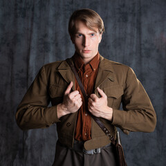 Fototapeta na wymiar A young guy in military-style clothes, a brown flight jacket and breeches with suspenders. Posing in the studio on a gray background, close-up