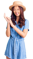 Fototapeta premium Young beautiful chinese girl wearing summer hat clapping and applauding happy and joyful, smiling proud hands together