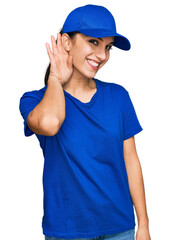 Young hispanic girl wearing delivery courier uniform smiling with hand over ear listening an hearing to rumor or gossip. deafness concept.
