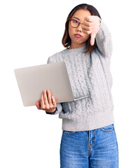 Young beautiful chinese girl wearing glasses holding laptop with angry face, negative sign showing dislike with thumbs down, rejection concept