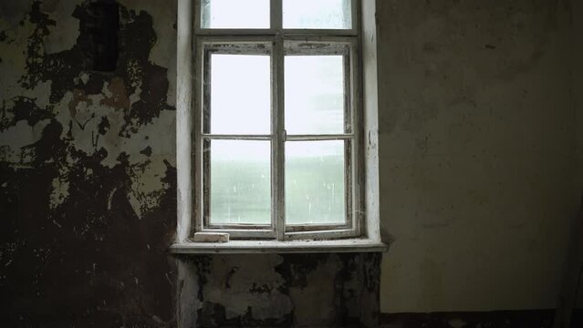 Tall old window with shabby and peeling paint on wooden frame in abandoned empty room, smooth approach of stabilized camera. Dirty window sill overgrown with cobwebs.