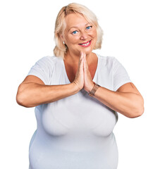 Middle age blonde woman wearing casual white t shirt praying with hands together asking for forgiveness smiling confident.