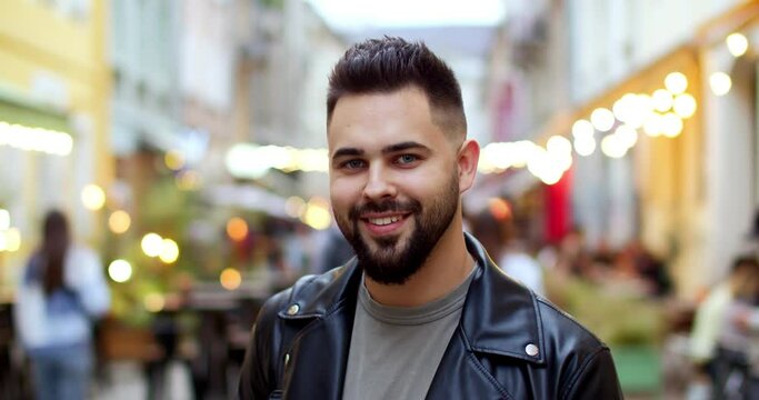 Portrait of young Caucasian handsome man with beard and in black leather jacket standing at decorated city street with lights on holiday. Close up. Decorations concept. Male in front of camera.