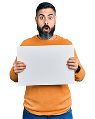 Hispanic man with beard holding blank empty banner afraid and shocked with surprise and amazed expression, fear and excited face.