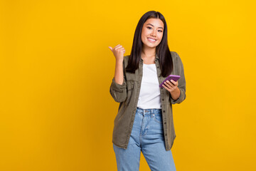 Photo of cheerful positive cute woman straight hairdo wear gray shirt hold phone directing empty space isolated on yellow color background