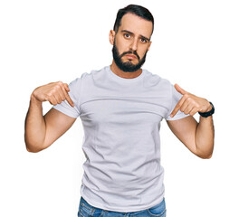 Young man with beard wearing casual white t shirt pointing down looking sad and upset, indicating...