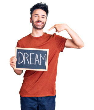 Young hispanic man holding blackboard with dream word pointing finger to one self smiling happy and proud