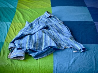 Wide shot. Blue, green and indigo linen shirt unfolded, dropped on a blue, green and indigo bed...