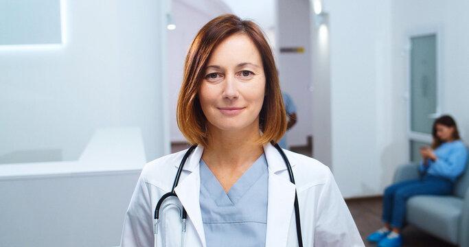 Close up portrait of happy Caucasian beautiful female physician with stethoscope in white coat standing in clinic, looking and smiling to camera. Healthcare concept